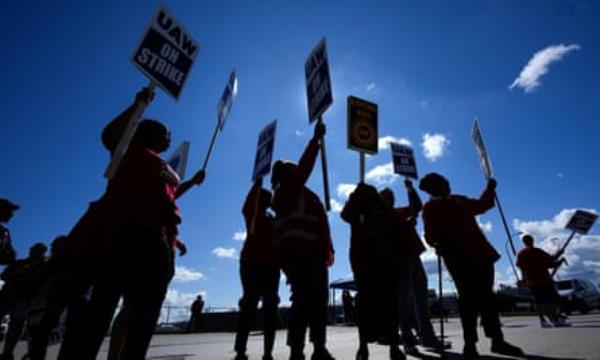 Taken from street level, people in silhouette beneath a sunny sky raise pickets signs.
